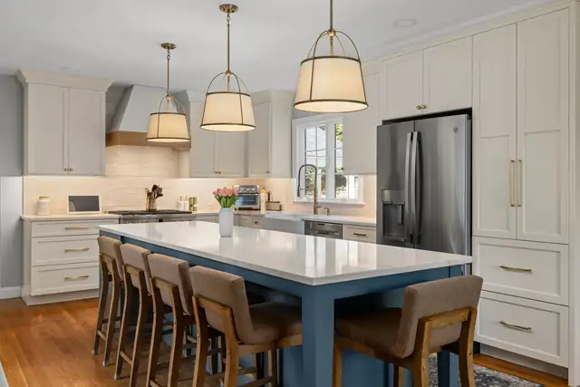 Kitchen by Transitions with cream shaker cabinets, a blue island with a white quartz top, wood counter stools and drum pendant lights
